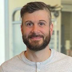A man with short brown hair and a beard, wearing a light gray henley shirt, smiles while standing indoors with blurred gym equipment on an urban campus in Toronto in the background.
