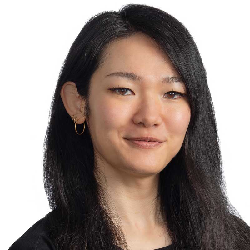 A woman with long, straight black hair and hoop earrings is smiling softly. She is wearing a black top and is posed against a plain white background.