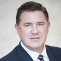 A man with short dark hair wearing a dark suit, white shirt, and striped tie, posing for a professional headshot against a light, blurred background.