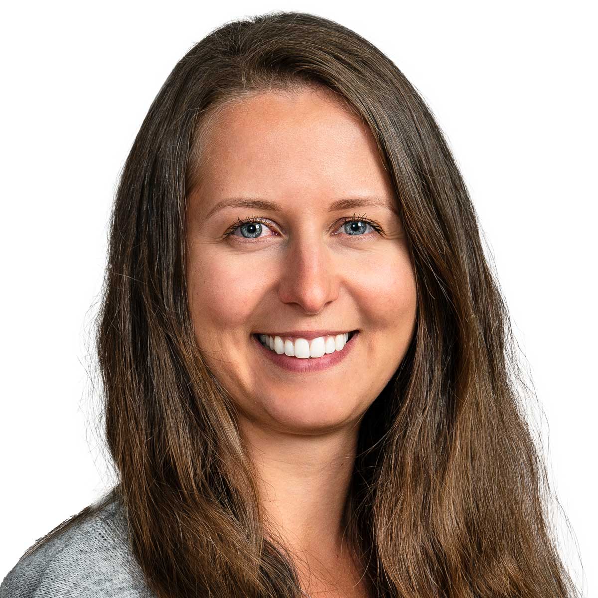 A woman with long brown hair and blue eyes smiles at the camera. She is wearing a light gray top and is posed against a plain white background.