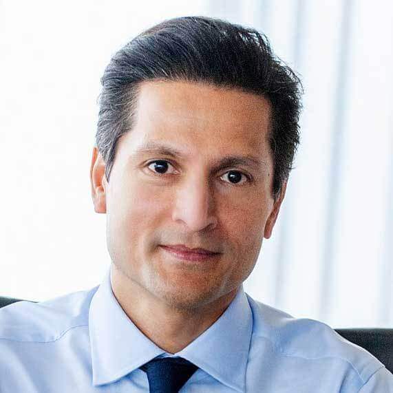 A man with short dark hair, wearing a light blue dress shirt and dark tie, sits indoors with a neutral expression; the softly blurred light background reflects the dynamic energy of an urban campus in Toronto.