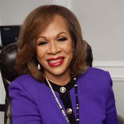A smiling woman with shoulder-length brown hair wears a purple blazer, black top, pearl necklace, and red lipstick, seated in an office chair with a light-colored background representing global education for girls.