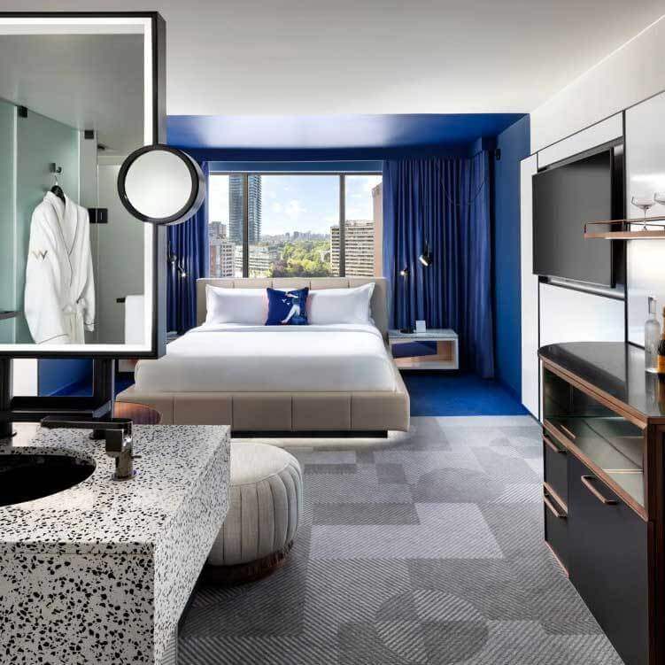 Modern hotel room with a white bed, blue accents, large window showcasing views of Toronto’s urban campus, contemporary bathroom with terrazzo countertop, mounted TV opposite the bed, and a white robe in the open bathroom area.