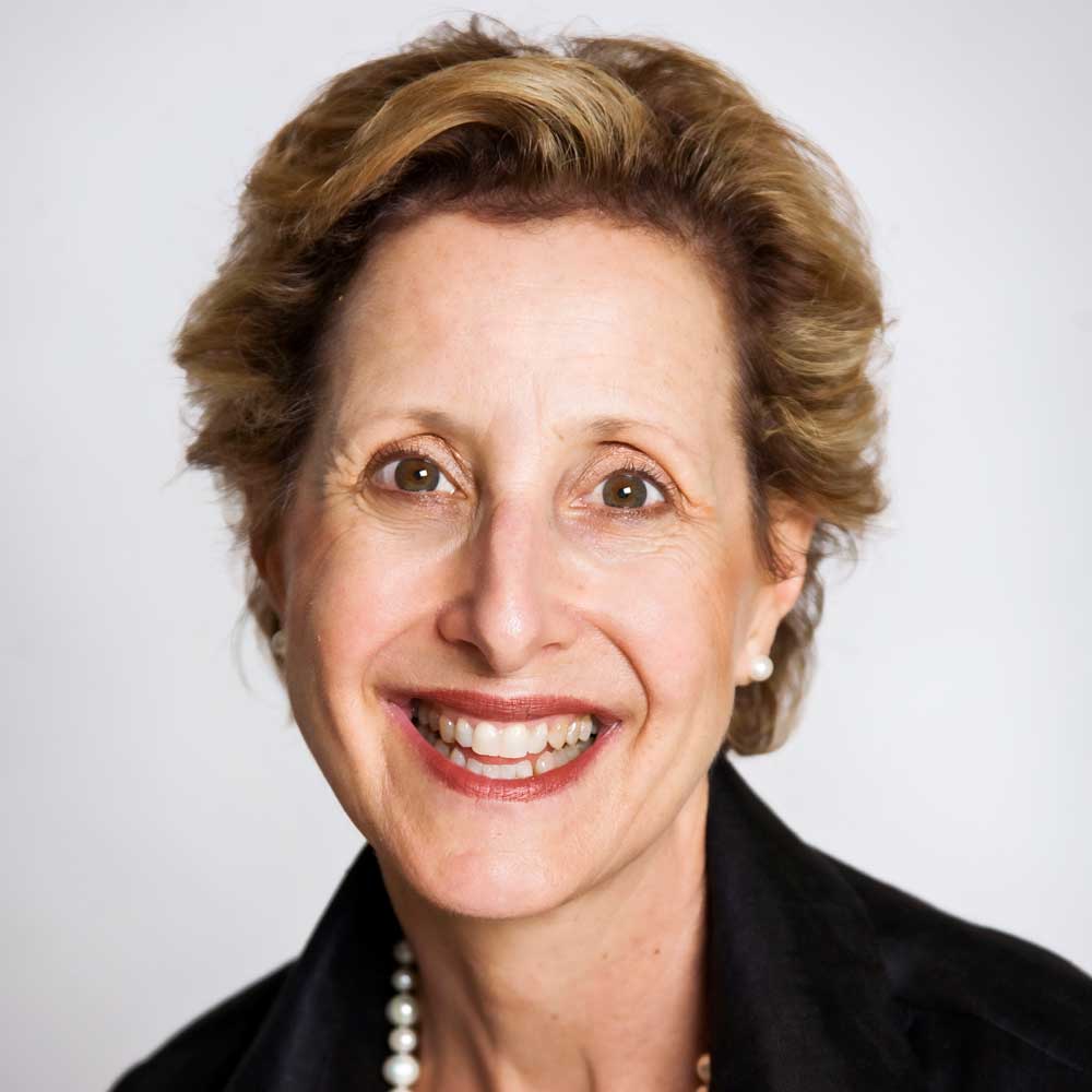 A smiling woman with short, light brown hair wearing a black top, a pearl necklace, and pearl earrings, posed against a plain light background.