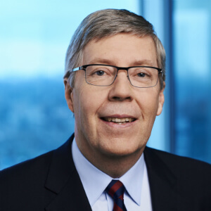 A middle-aged man with short gray hair, glasses, and a suit with a striped tie, smiling in an office setting with large windows and a blurred cityscape in the background.