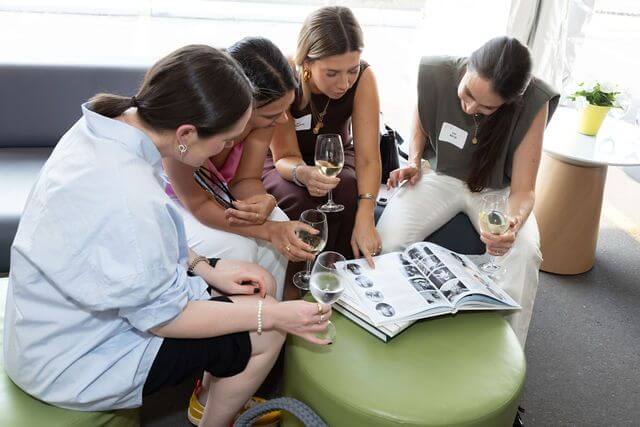 Four women sit together on ottomans, each holding a glass of white wine, engaged as they explore a magazine about global education for girls and scholarships for girls. Their focus suggests genuine interest in the opportunities featured within its pages.