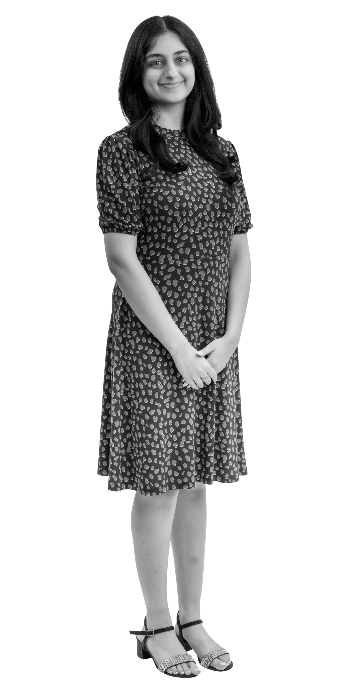 A woman with long dark hair stands and smiles, wearing a patterned knee-length dress and sandals. She has her hands clasped in front of her. The image is in black and white with a white outline.