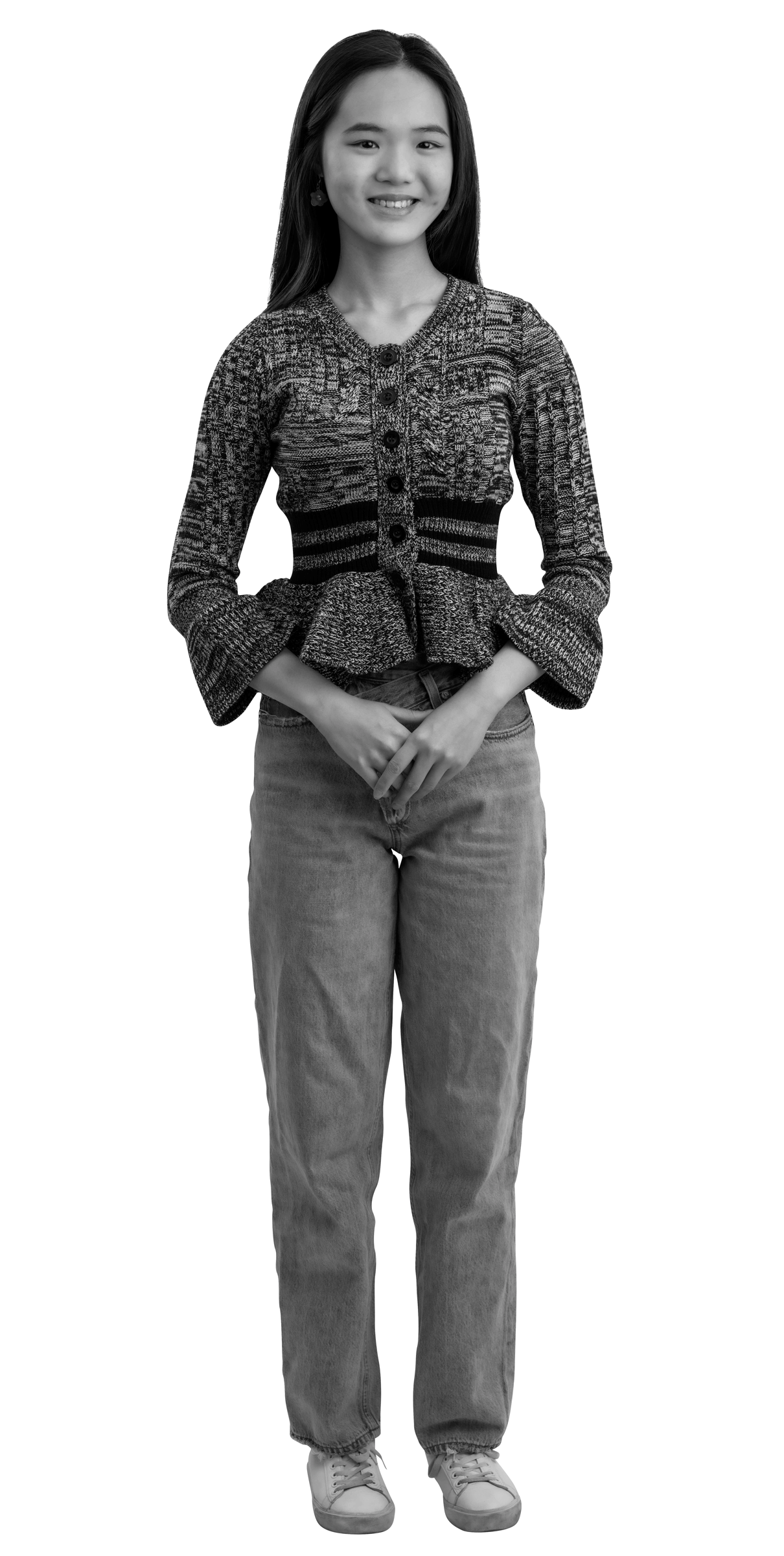 A young woman with long hair stands smiling, wearing a knitted sweater, jeans, and sneakers. Her hands are clasped in front of her. The image is in black and white with a white outline around her.