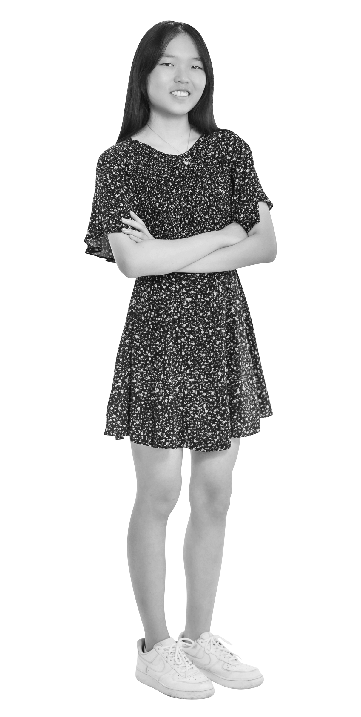 A young girl with long hair stands with arms crossed, wearing a short-sleeved, patterned dress and sneakers. She is facing forward and smiling slightly. The image is in black and white with a white outline around her.