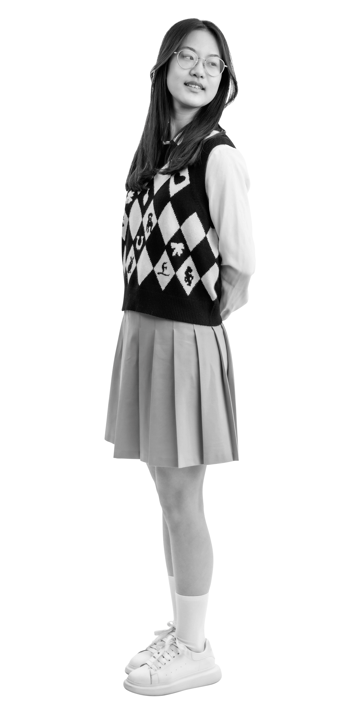A young woman wearing glasses, a patterned sweater vest over a collared shirt, a pleated skirt, knee-high socks, and sneakers stands with her hands behind her back, looking to the side and smiling.