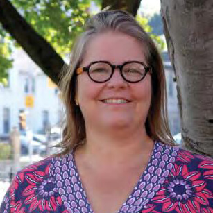 A woman with long, light brown hair and round glasses smiles outdoors. She is wearing a patterned shirt with pink and purple designs and is standing next to a tree. Green leaves and a blurred building are in the background.