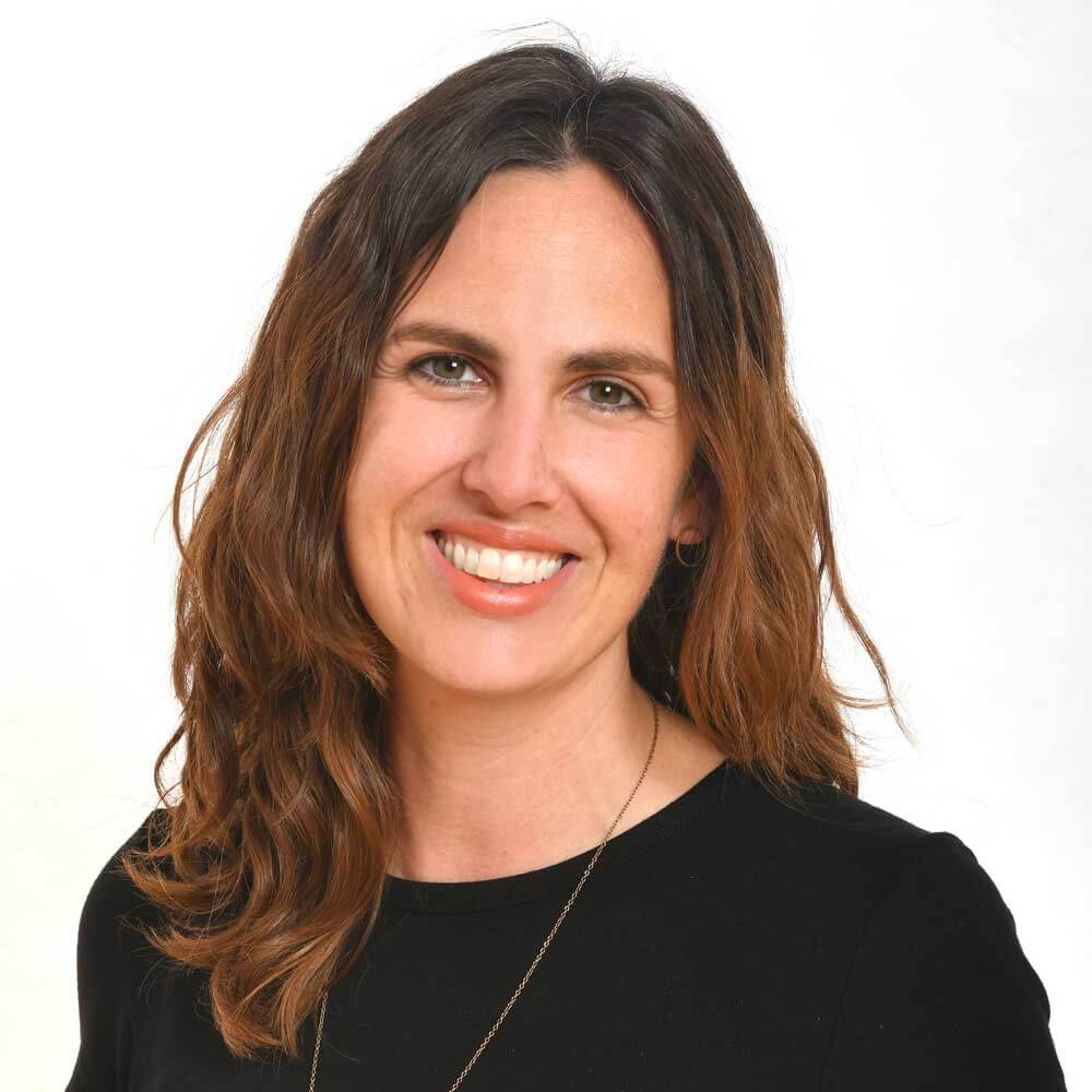 A woman with wavy brown hair and a friendly smile, wearing a black top and a necklace, poses in front of a plain white background.