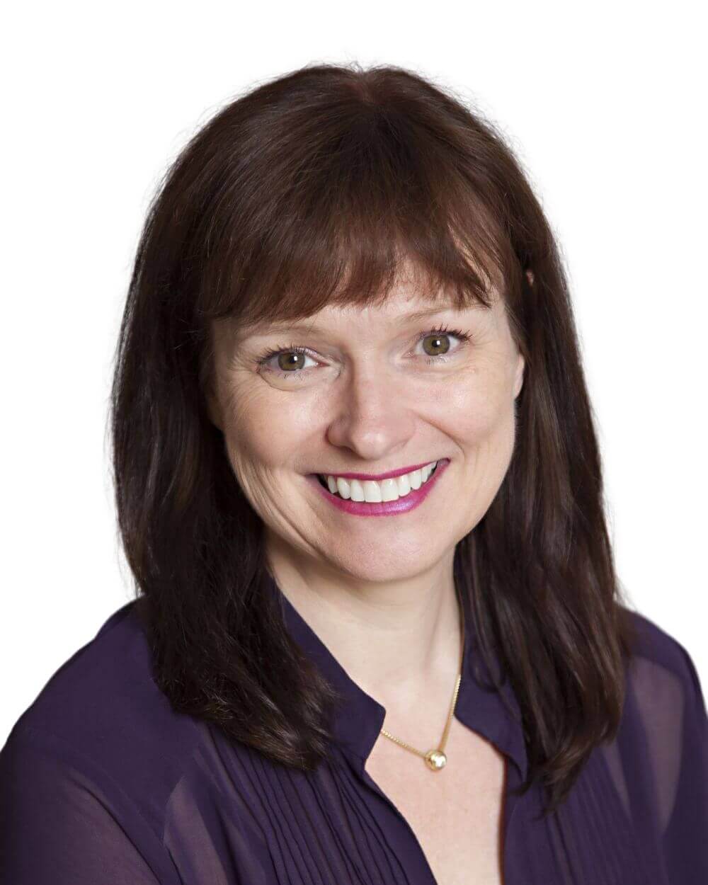A woman with straight brown hair and bangs smiles at the camera. She is wearing a dark purple blouse and a gold necklace with a small pendant. The background is plain white.