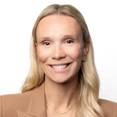 A woman with long blonde hair, wearing a brown blazer and a delicate necklace, smiles at the camera against a plain white background.