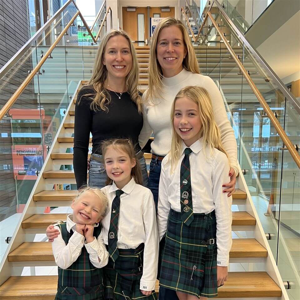 Two women stand on stairs behind three young girls wearing matching green plaid skirts and ties. Everyone is smiling. The group appears to be in a school building with glass railings and wooden steps.