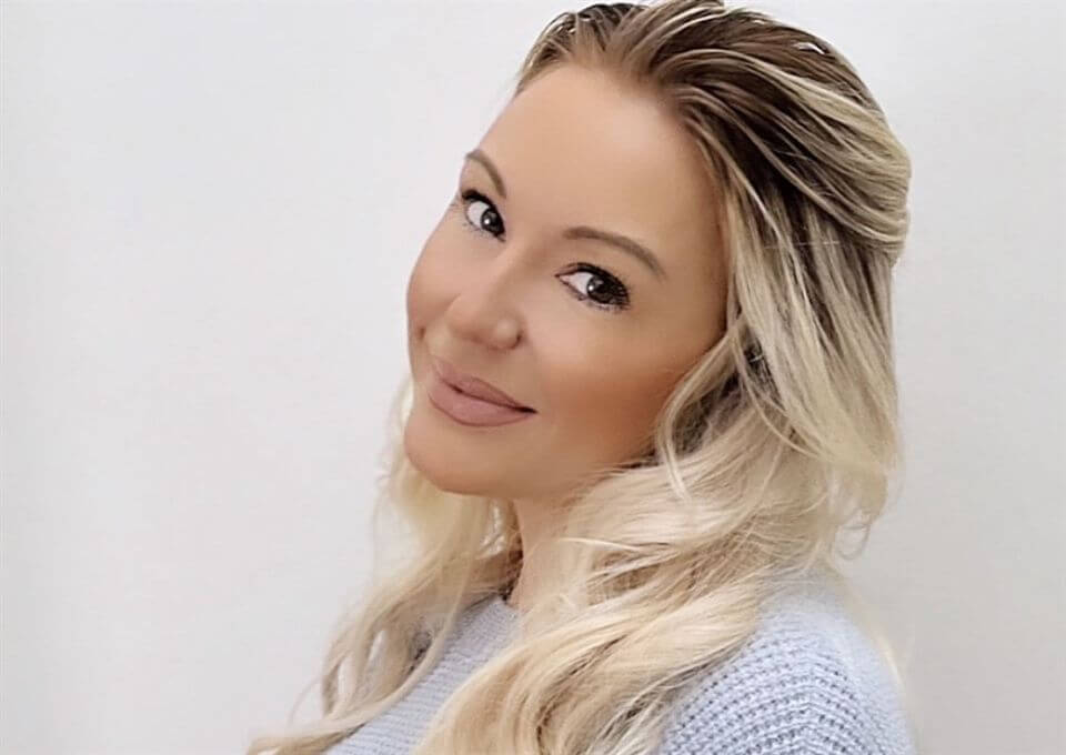 A woman with long, wavy blonde hair and light makeup smiles softly, tilting her head slightly to the side against a plain white background. She is wearing a light blue knit sweater.