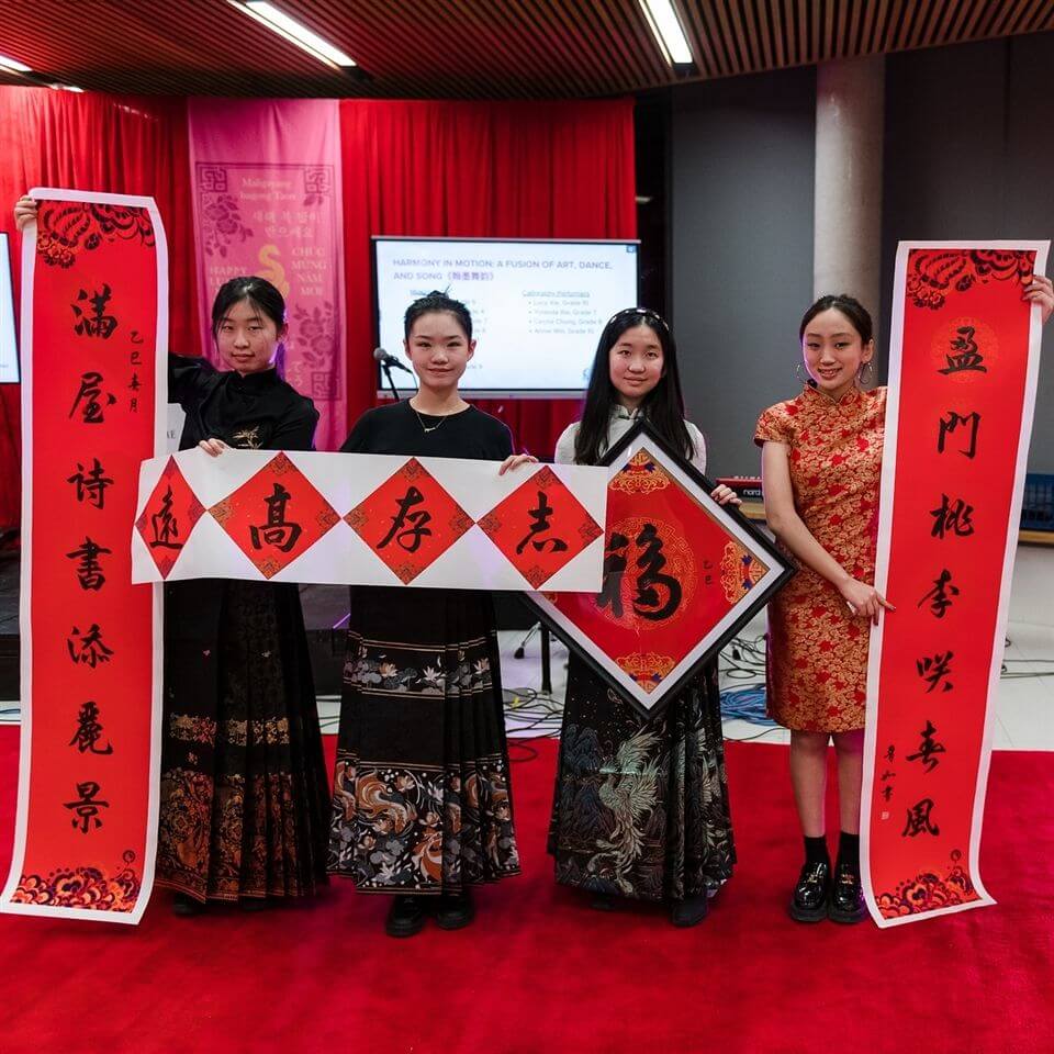 Four young women dressed in traditional Chinese attire stand on a red carpet, each holding large red and white calligraphy banners with Chinese characters. Red curtains and decorations are visible in the background.