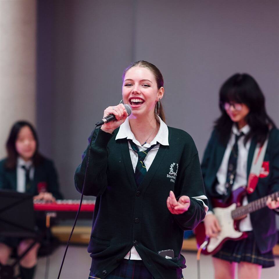 A smiling teenage girl in a school uniform sings into a microphone while two other students play keyboard and guitar in the background, all wearing matching green blazers and plaid skirts.