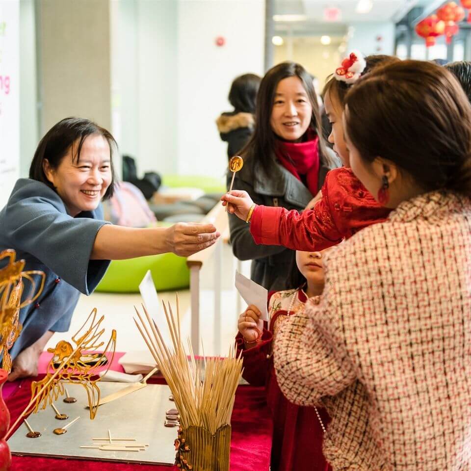 A woman smiles as she hands a sugar sculpture on a stick to a child across a table decorated with dragon-shaped sugar art. Several people, including children, stand around the table in a festive indoor setting.