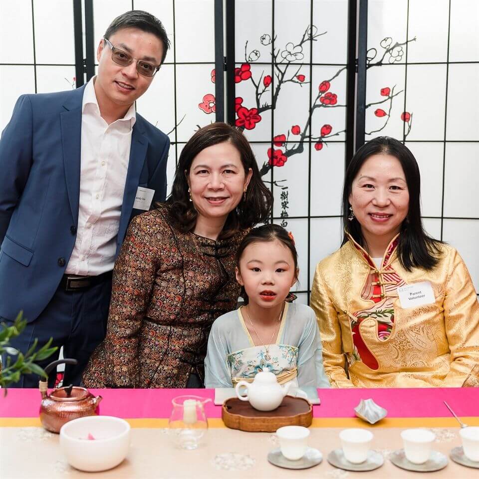 Four people, including a young girl in traditional clothing, pose together at a tea ceremony table. The backdrop features a decorative screen with red flowers and branches. Tea cups and a pot are on the table in front of them.