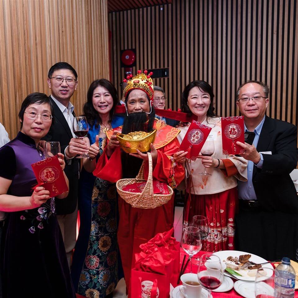 A group of six adults, including a person dressed as the Chinese God of Wealth, smile and hold red envelopes at a festive indoor gathering. Wine glasses and plates are on the table in front of them.