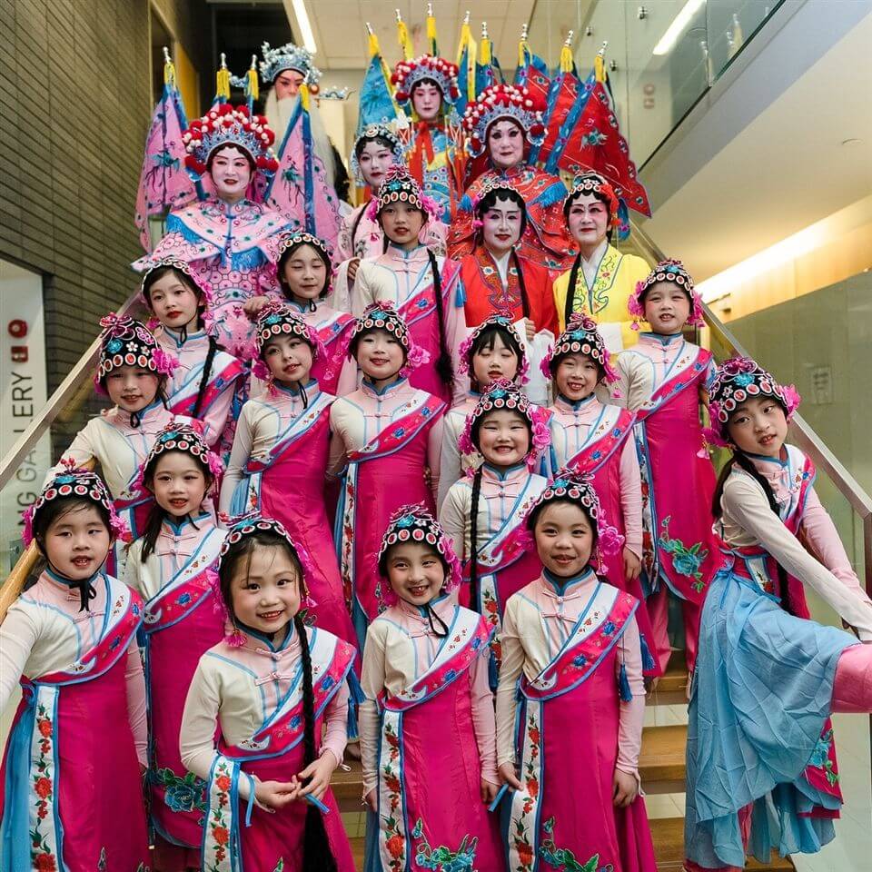 A group of children and adults in bright pink, blue, and red traditional Chinese opera costumes pose together on a staircase, smiling for the camera. The costumes feature ornate headpieces and colorful patterns.