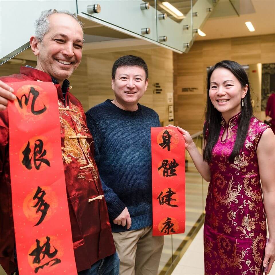 Three people smile and hold red banners with Chinese calligraphy, celebrating indoors. One man wears a red patterned shirt, another wears a blue sweater, and a woman wears a red cheongsam. The setting appears festive and joyful.