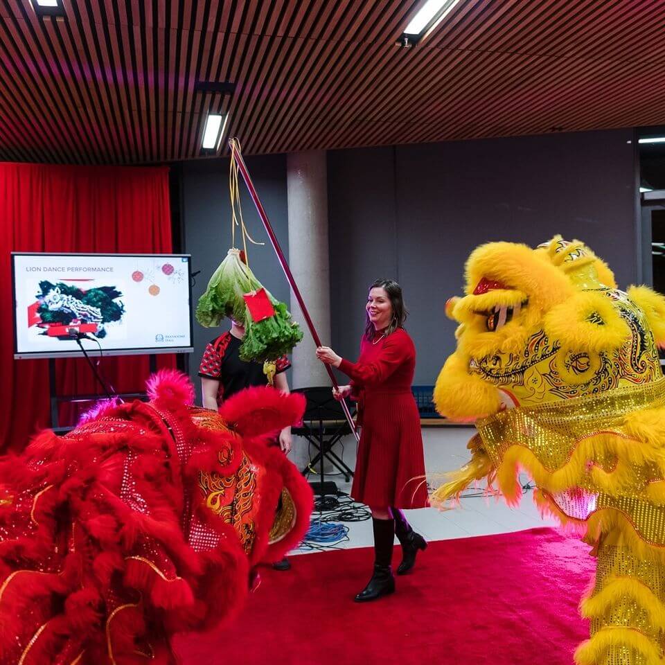 A woman in a red dress holds a pole with greens and red envelopes as two vibrant lion dance costumes—one red, one yellow—approach her on a red carpet, with a presentation about lion dance visible on a screen in the background.