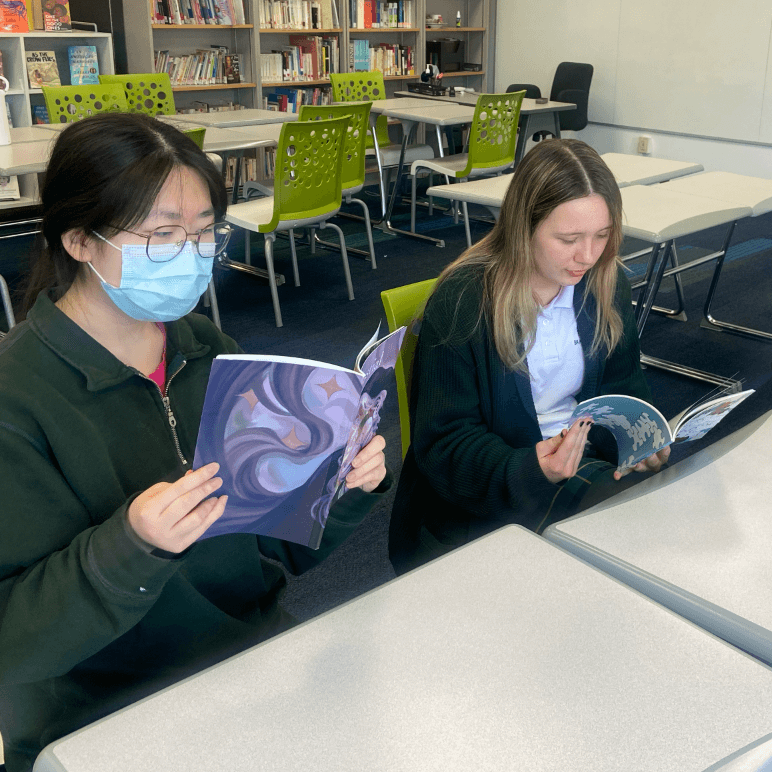Two students sit at desks in a library, reading graphic novels. One wears glasses and a blue face mask; both are focused on their books. Bookshelves and green chairs are visible in the background.
