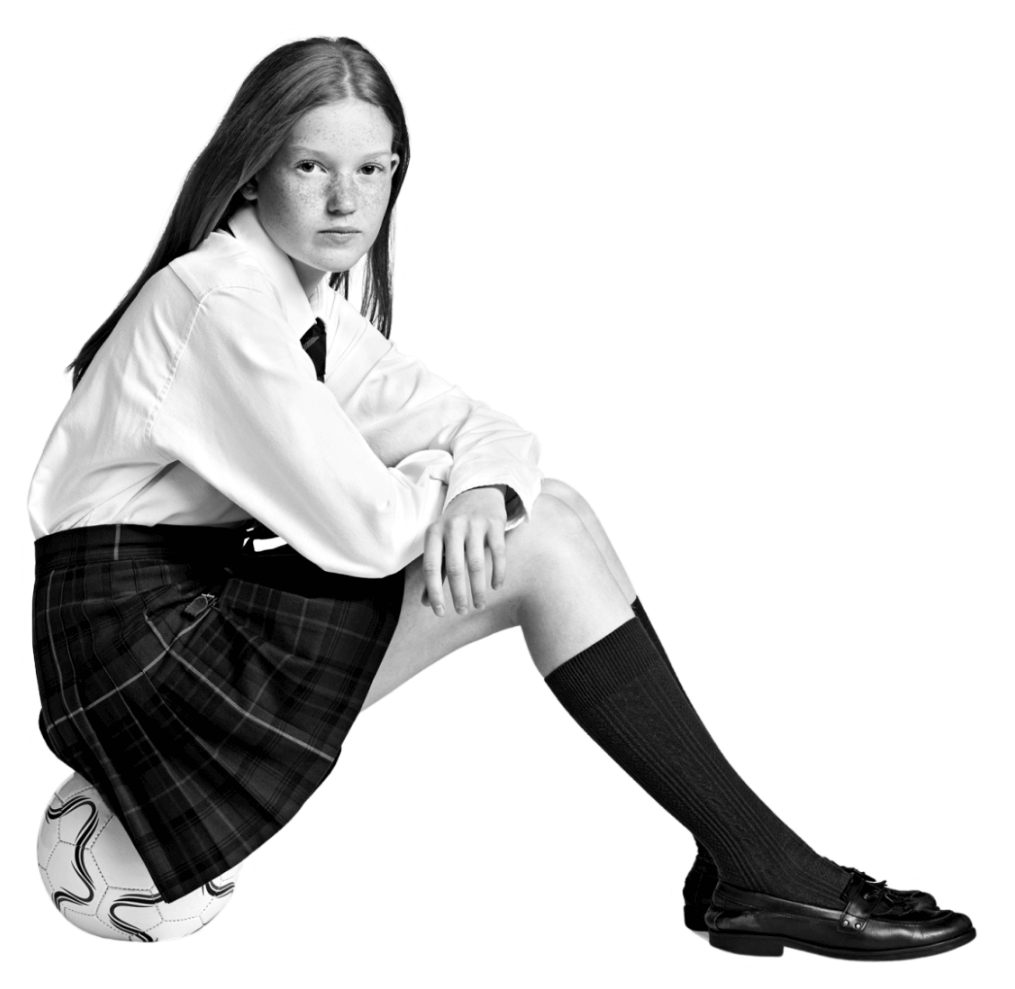 A young person with long hair, wearing a white shirt, plaid skirt, tie, and knee-high socks, sits on a soccer ball with arms resting on their knees. The image is in black and white with a plain background.