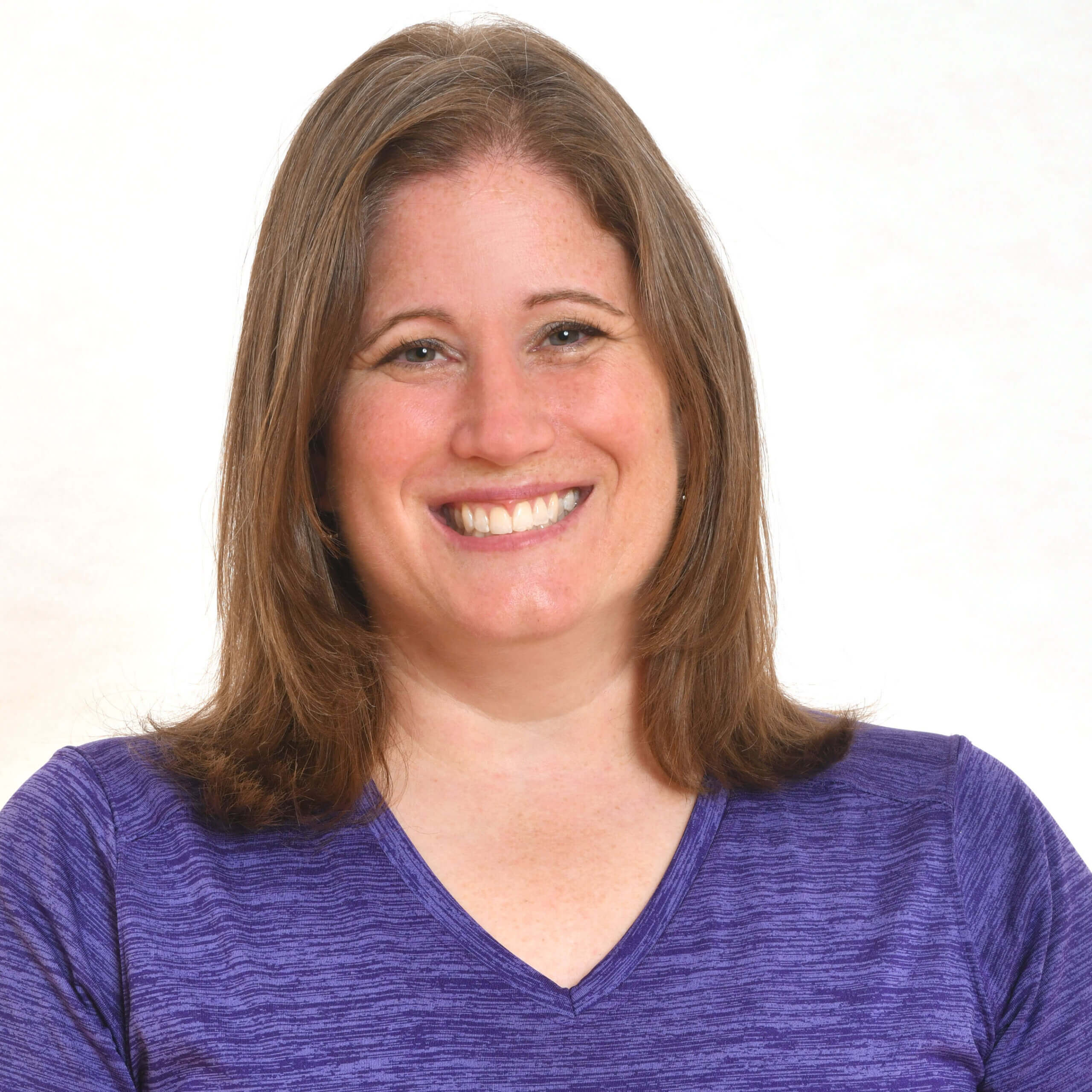 A woman with straight, shoulder-length light brown hair smiles at the camera. She is wearing a purple V-neck shirt and is posed in front of a plain, light background.