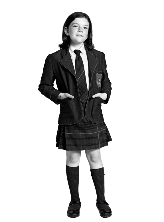 A young student in a school uniform, including a blazer, tie, plaid skirt, long socks, and dress shoes, stands with hands in blazer pockets on an urban campus Toronto—confidently representing global education for girls. The image is black and white.