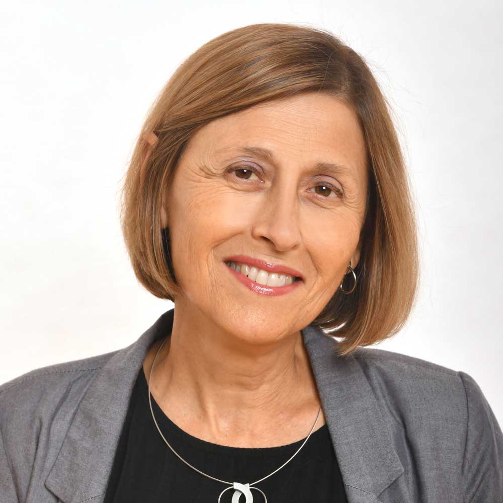 A woman with short, straight light brown hair is smiling. She is wearing a gray blazer, a black top, hoop earrings, and a necklace, with a plain white background.