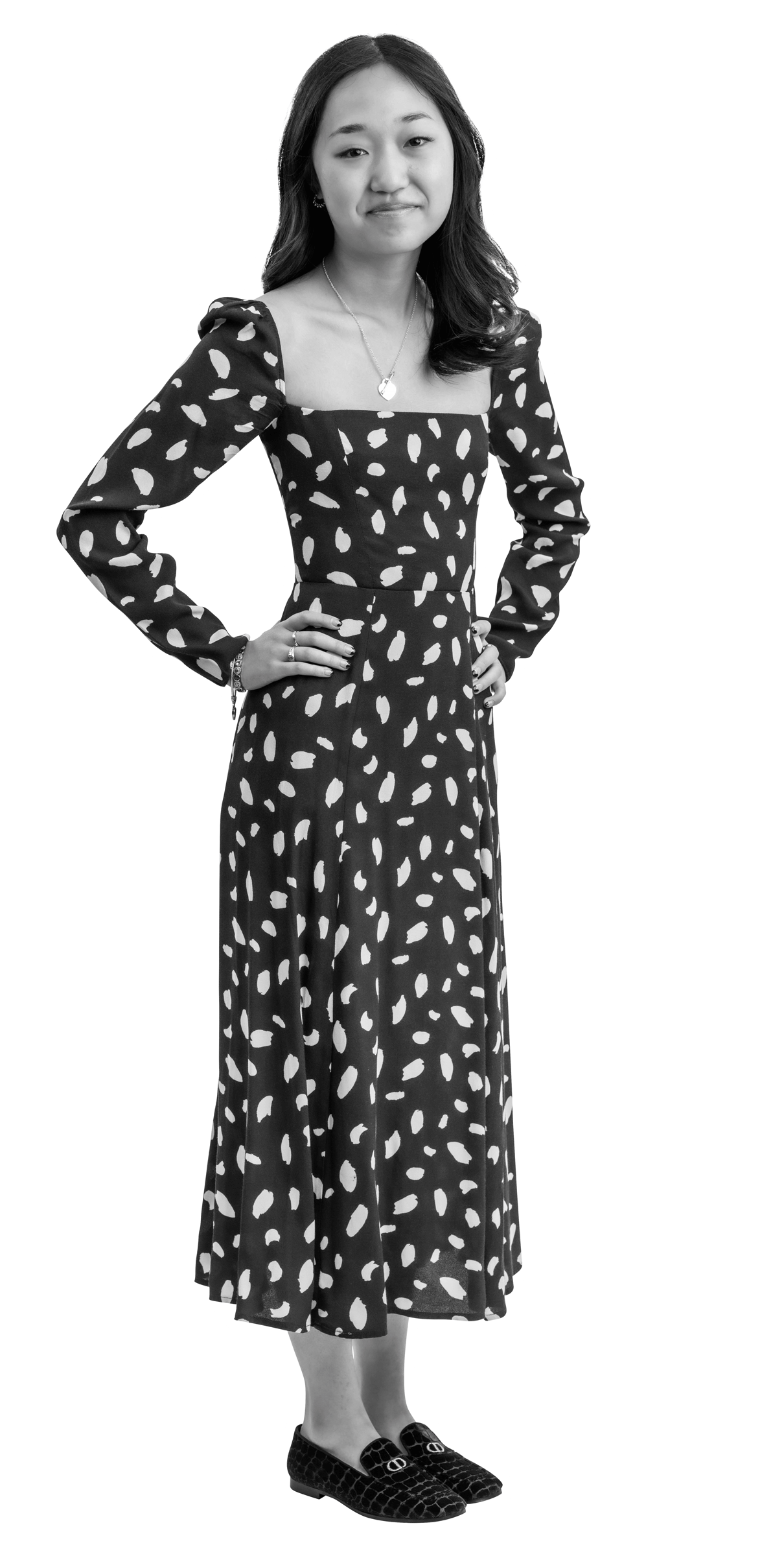 A young woman stands with hands on hips, wearing a long, patterned dress with a square neckline and dark loafers. She has long, wavy hair and a neutral expression. The image is in black and white with a white outline.