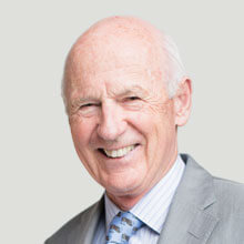 An older man with a bald head smiles warmly. He is wearing a light gray suit, a blue striped shirt, and a patterned tie, with a plain light background.