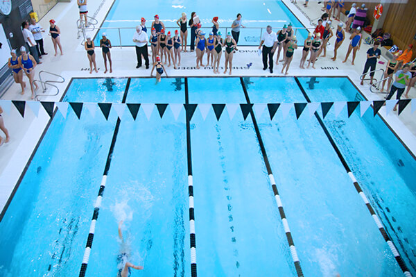 Aerial view of an indoor swimming pool during a swim meet, with swimmers racing in lanes and spectators and officials standing along the poolside.