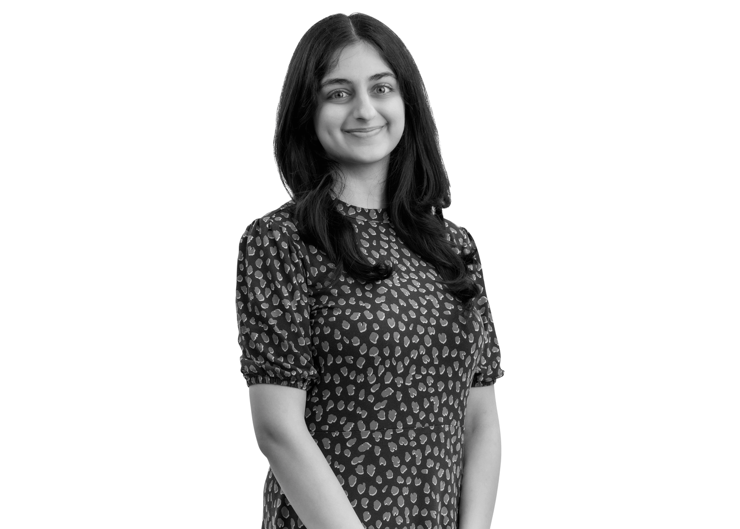 A woman with long dark hair wearing a patterned dress stands smiling, with her hands clasped in front of her, against a plain white background.