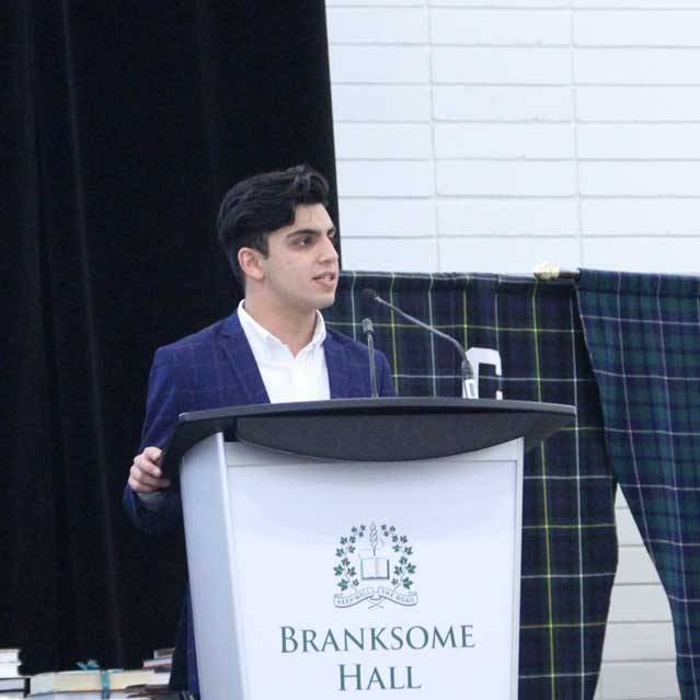 A young man in a blue suit speaks at a podium labeled "Branksome Hall," with a microphone and dark curtain behind him.