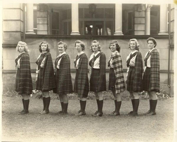 Eight young women stand in a row outside a building, wearing plaid skirts, capes, and knee-high socks, posing for a formal group photo in front of a large stone structure with columns.