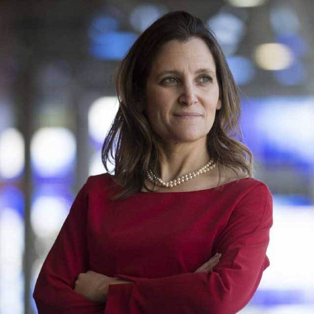 A woman with long brown hair, wearing a red dress and a pearl necklace, stands with arms crossed and looks to the side, with a blurred background of blue and white lights.