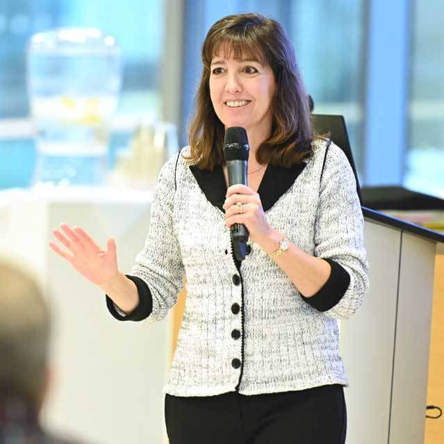 A woman with shoulder-length brown hair speaks into a microphone, smiling and gesturing with her left hand. She is wearing a light sweater and standing indoors near a podium with a water dispenser in the background.