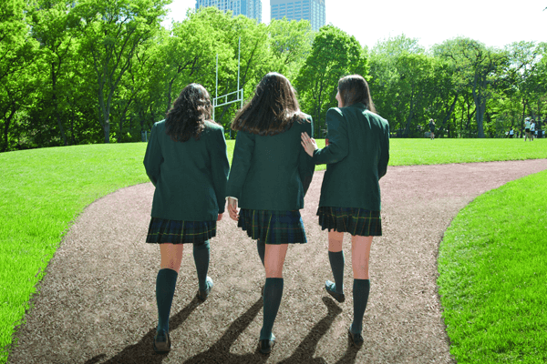 Three students in green blazers and plaid skirts walk together on a path across a grassy field, with trees and tall buildings visible in the background.