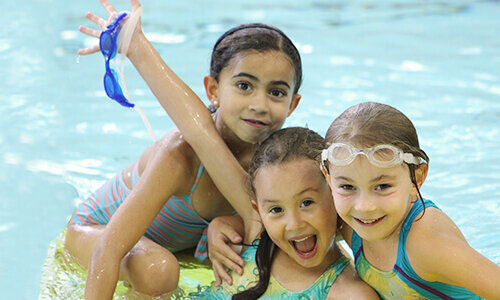 Three young girls in swimsuits smile and pose together in a swimming pool; one girl holds up a pair of blue goggles while the others wear goggles on their heads.
