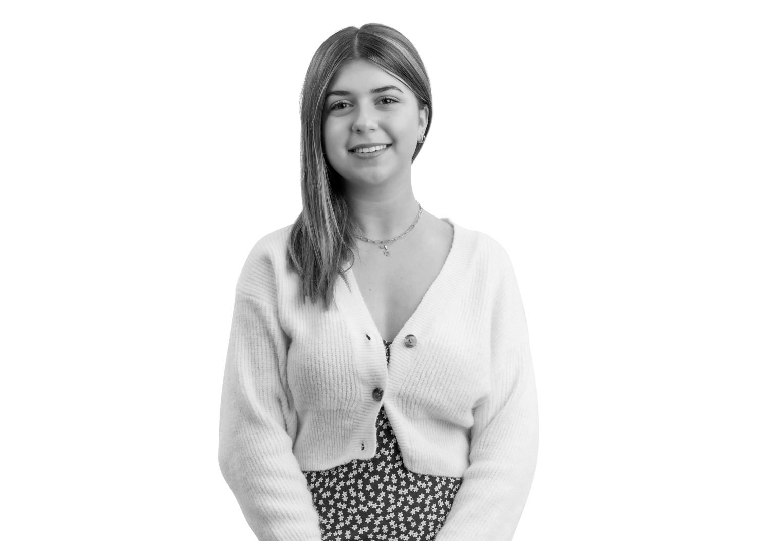 A young woman with straight, shoulder-length hair smiles at the camera. She is wearing a light-colored cardigan over a patterned dress and a necklace, standing against a plain white background.