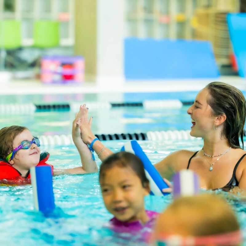 A swim instructor smiles and high-fives a young girl wearing goggles and a red life vest in a swimming pool, while other children swim nearby.
