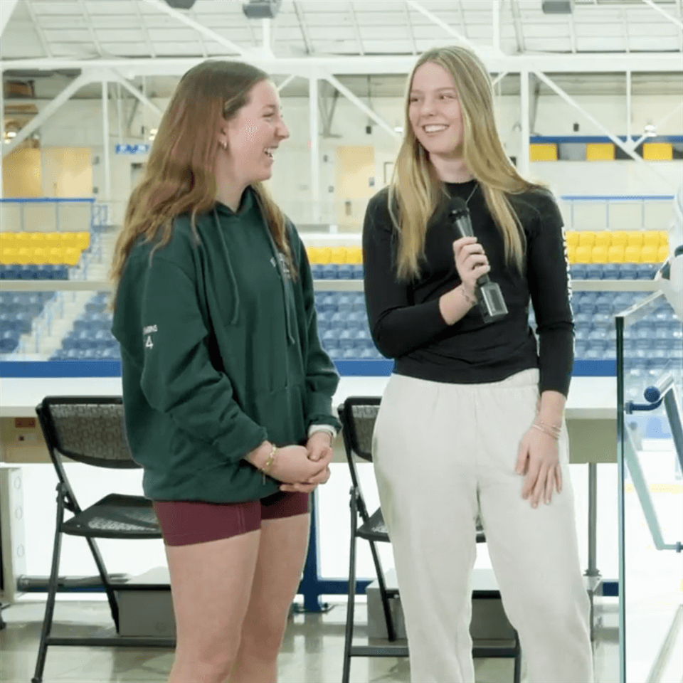 Two young women stand and smile at each other in an indoor arena. One holds a microphone, wearing a black top and white pants. The other wears a green hoodie and maroon shorts. Rows of blue and yellow seats are visible behind them.