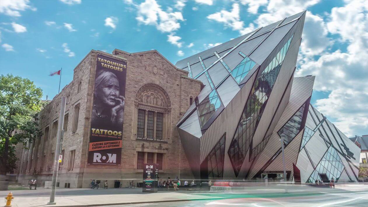 The Royal Ontario Museum in Toronto, featuring its historic stone building beside the modern, angular glass Crystal Wing addition, with a street view and a large "Tattoos" exhibition banner on the facade.