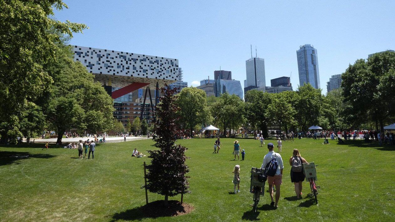 People walk and relax on a large grassy park surrounded by trees, with a modern building featuring a checkerboard design and tall city skyscrapers in the background under a clear blue sky.