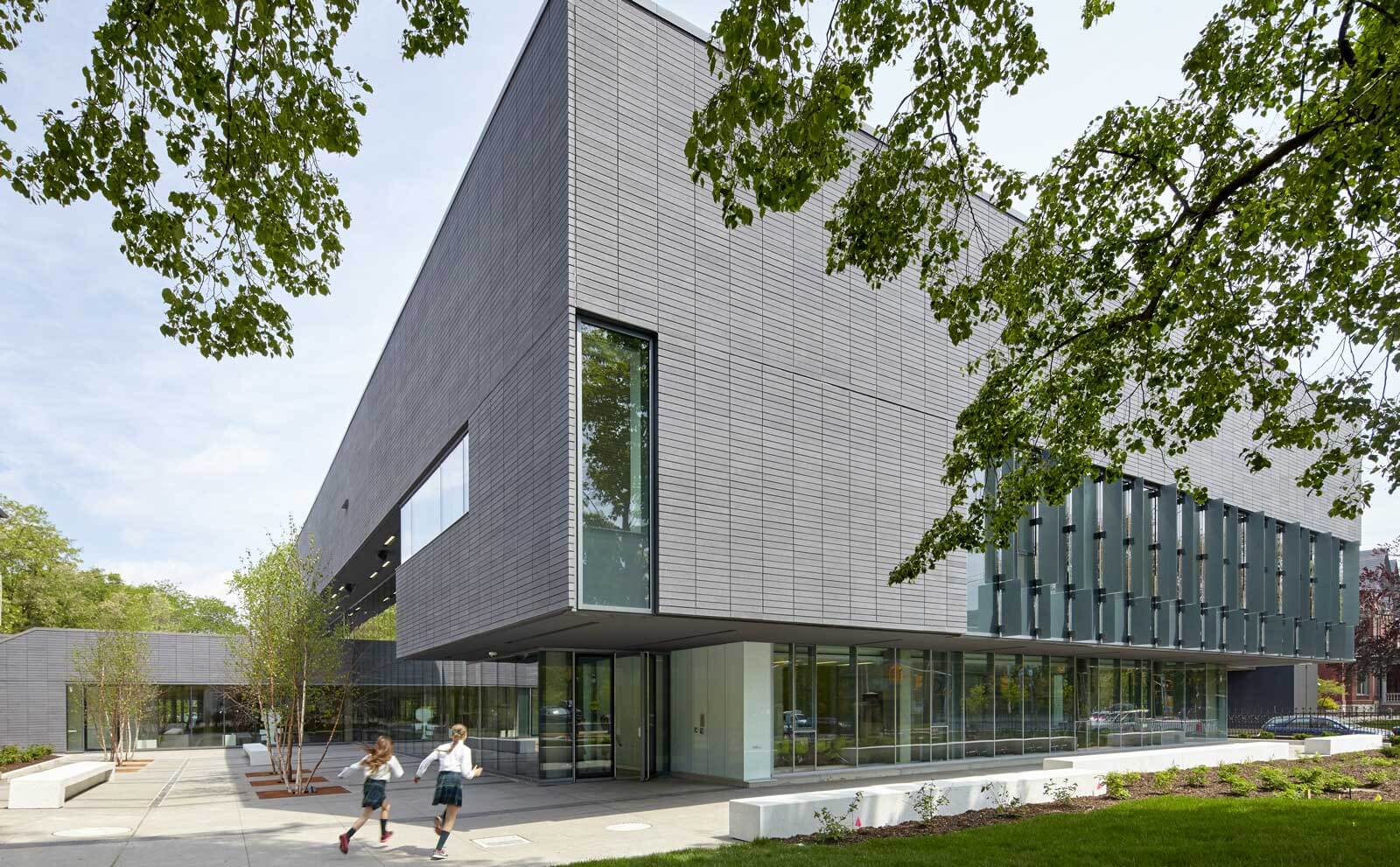 Two children run on a sidewalk outside a modern building with large glass windows and gray horizontal paneling, surrounded by trees and landscaped greenery.