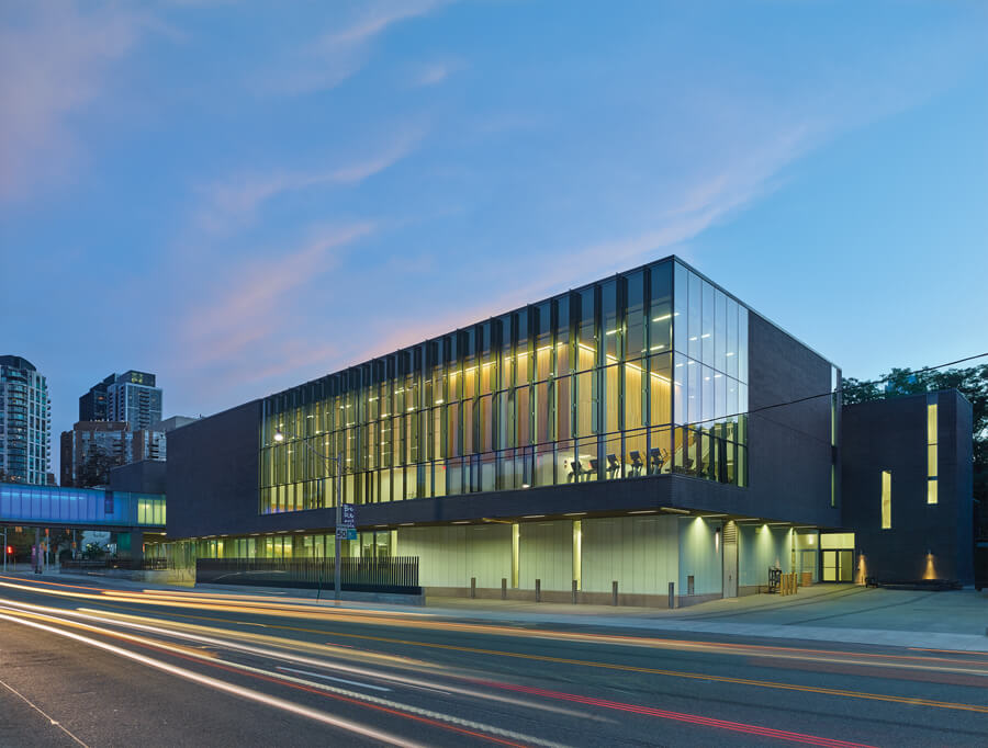 Modern glass and brick building at dusk, with large windows glowing from interior lights. Light trails from passing cars line the street in front. City buildings and a sky bridge are visible in the background.